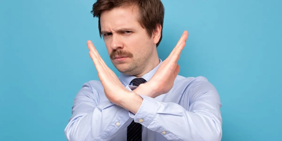 serious young man in shirt showing stop sign crossing hands and looking at camera isolated blue background