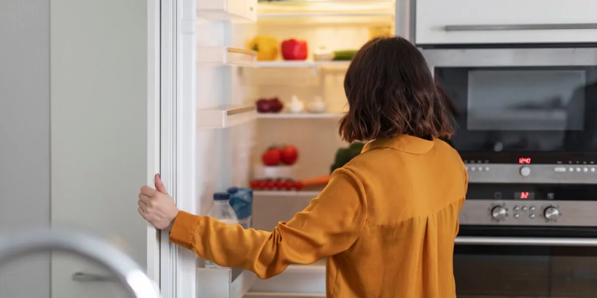 unrecognizable young woman opening fridge in kitchen and looking inside, hungry brunette lady standing near open refrigerator at home, starving female checking food or drinks, free space