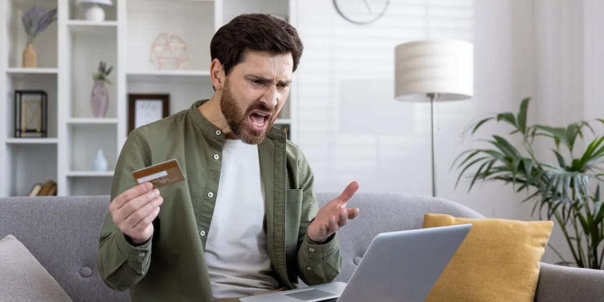 man expressing frustration with online payment holding credit card and using laptop on couch at home concept of online shopping issues, payment failure, and internet banking problems