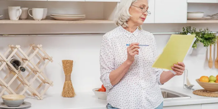pensive senior housewife is signing insurance contract she is standing in domestic kitchen