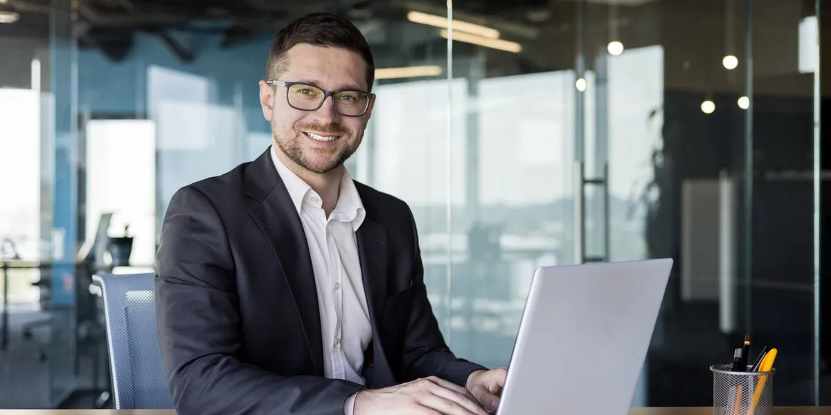 portrait of a successful male businessman, banker, financial director sitting in the office at the table and using a laptop looking and smiling at the camera
