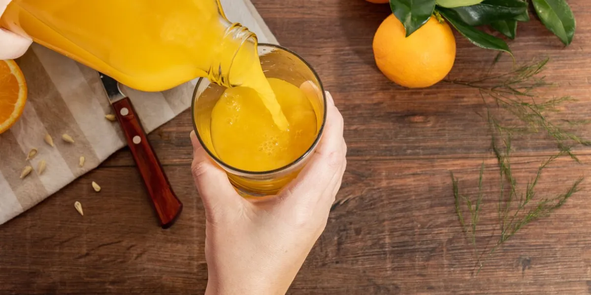 a person pouring orange juice into a glass cup in the background you can see whole oranges and a knife resting on a wooden table