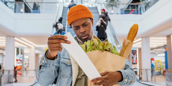 surprised african-american man in denim jacket looks at receipt total in sales check holding paper bag with products in mall