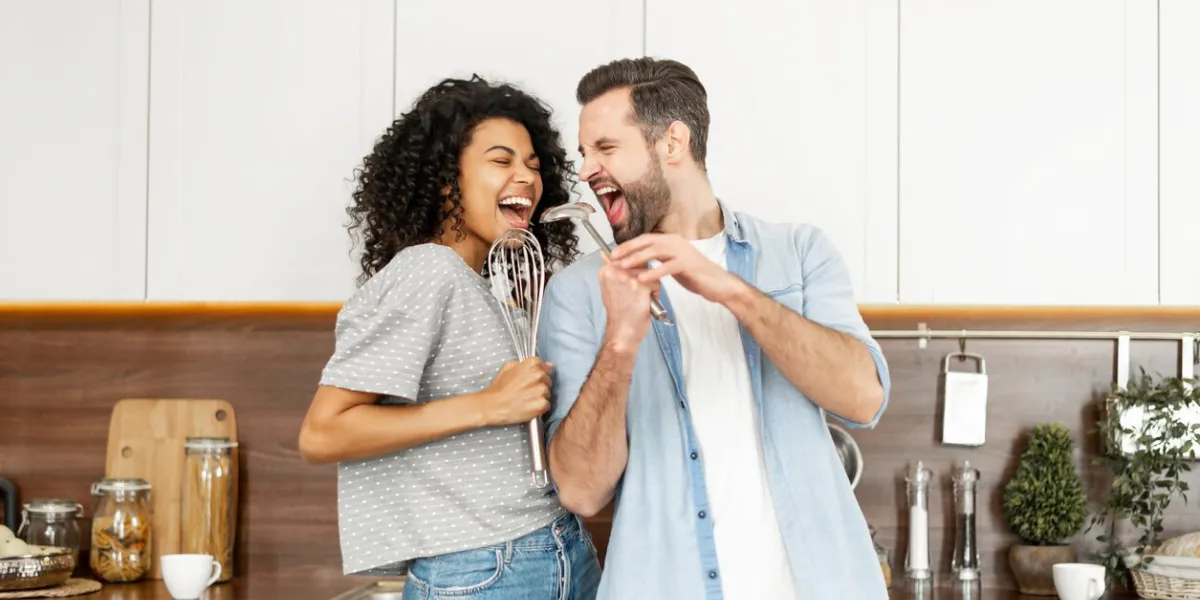 happy interracial couple dancing in the kitchen, singing while cooking breakfast or dinner, young african american woman and a handsome man smiling and having fun together, holding kitchen utensils