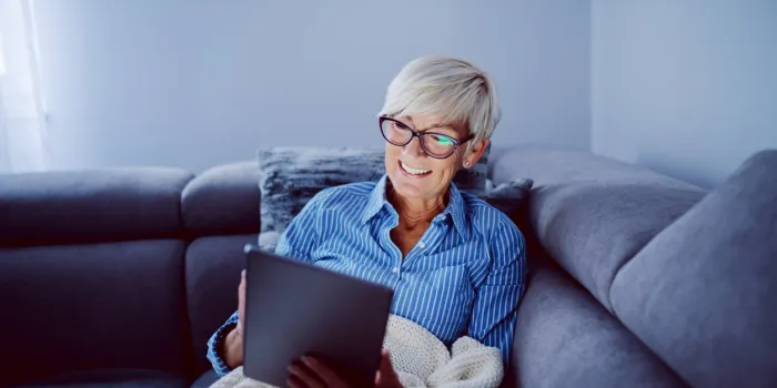 beautiful smiling blonde caucasian senior woman sitting on sofa in living room and using tablet for internet surfing