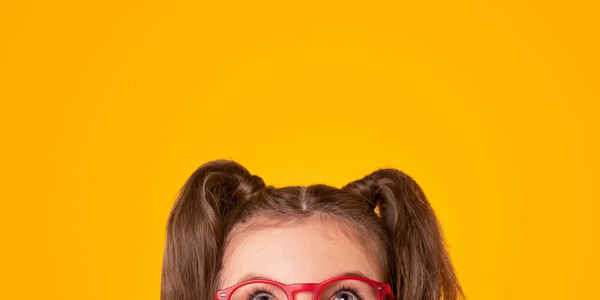 positive teen girl with ponytails wearing stylish red framed eyeglasses looking up on yellow background with empty space